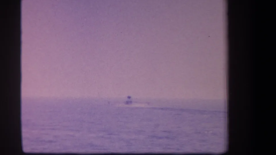 1959 8mm frame showing a small distant aircraft near the horizon over open water, with a lavender dusk cast.