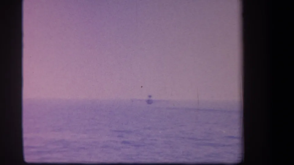 1959 8mm frame showing the aircraft as a small silhouette over water, with soft focus and visible film aging.