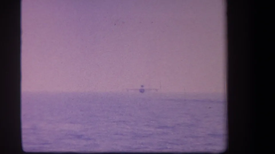 1959 8mm frame with a distant plane near the water horizon, framed by the soft faded color of aged film.