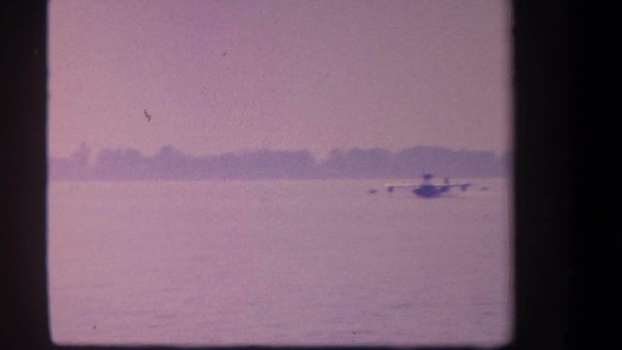 Seaplane skims low over the lake with a hazy treeline in the distance.