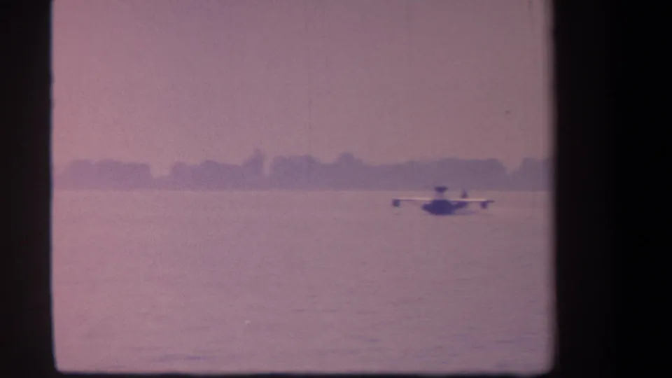 1959 8mm frame of a seaplane on or just above the lake surface, with a blurred treeline beyond the water.