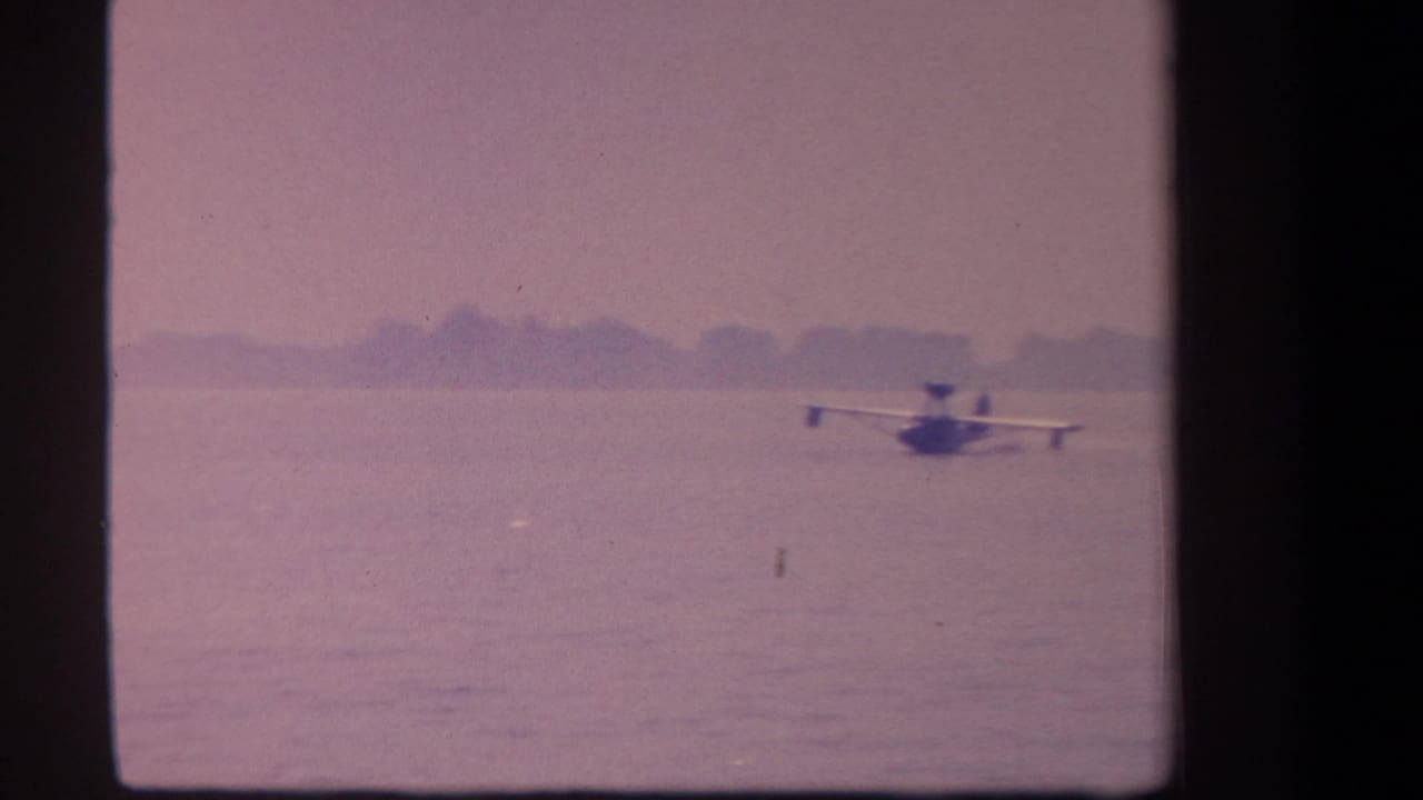 1959 8mm frame from California metadata showing a small floatplane on calm water under a faded purple sky.