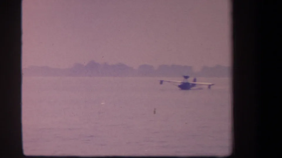 1959 8mm frame from California metadata showing a small floatplane on calm water under a faded purple sky.