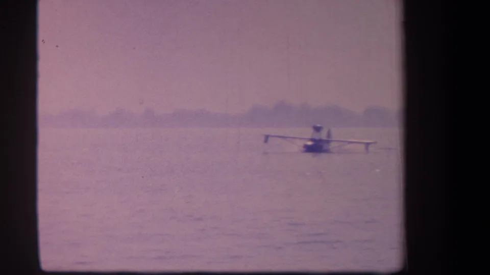 A small floatplane appears on the water in a faded 1959 8mm frame, with shoreline trees in the distance.