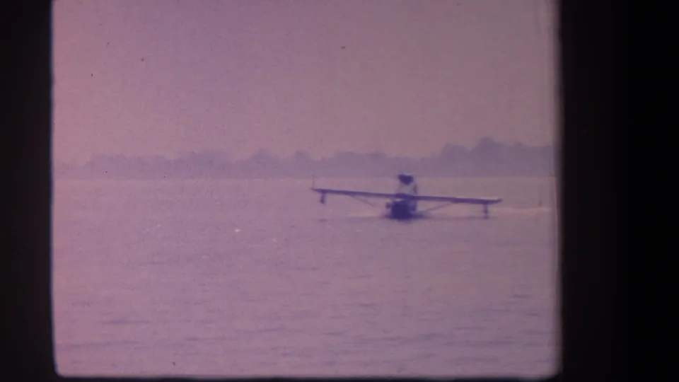 The floatplane sits near center frame on calm water, captured in soft, purple-toned 8mm home-movie footage.