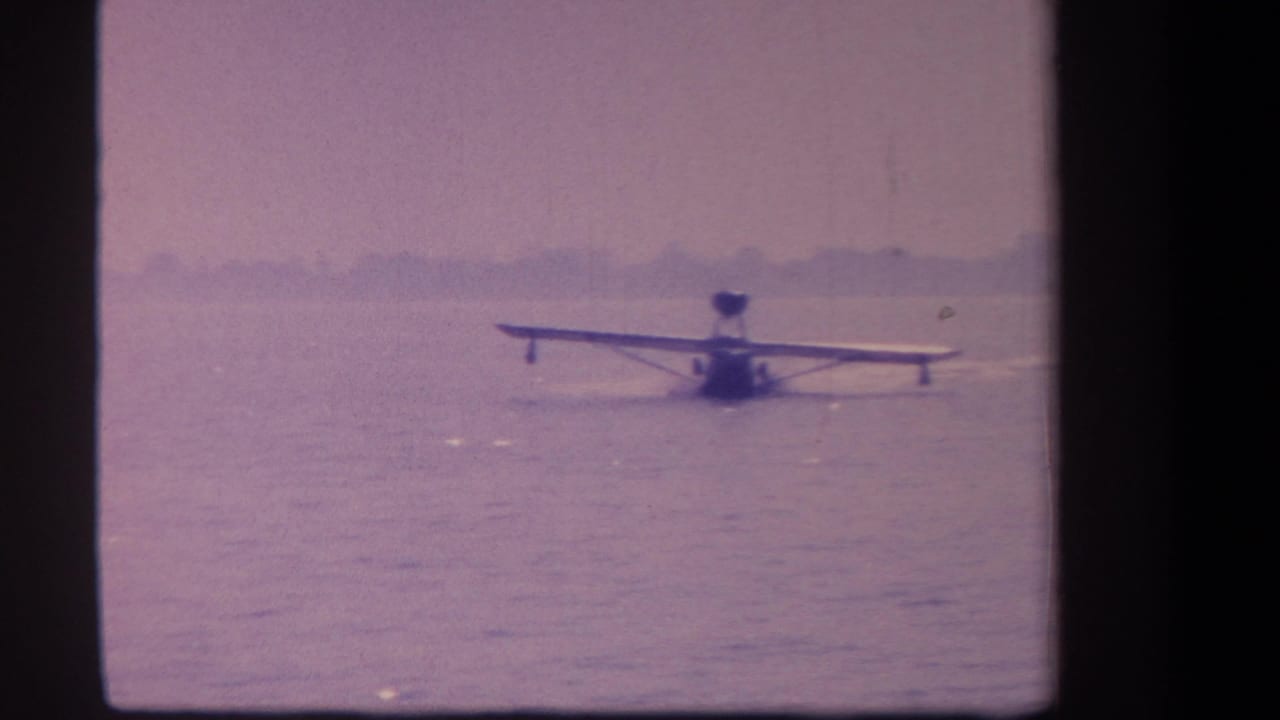 1959 California 8mm frame showing a small float plane on calm water, seen from a distant shore position.