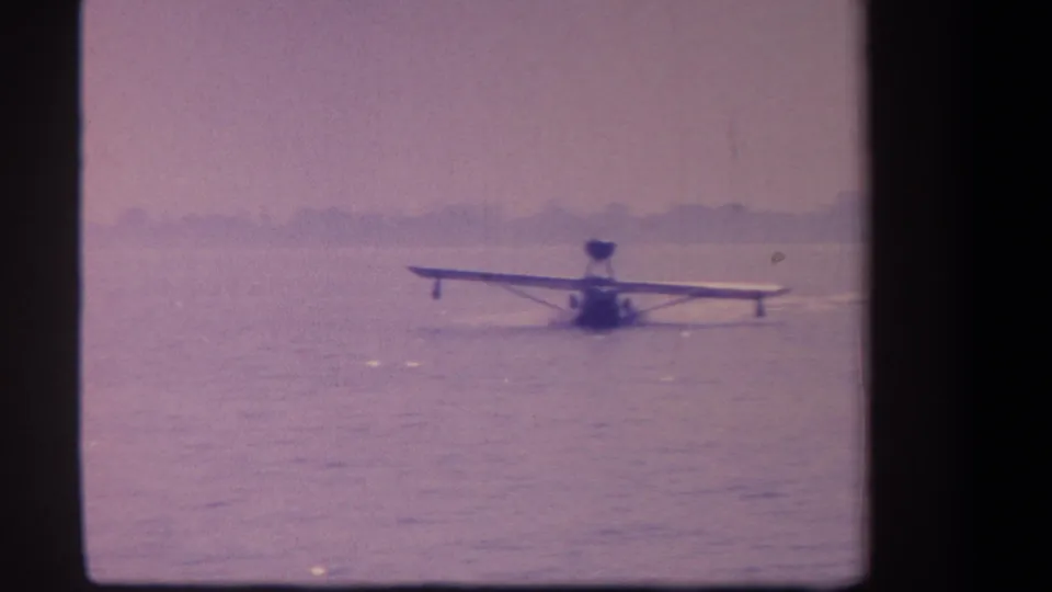 1959 California 8mm frame showing a small float plane on calm water, seen from a distant shore position.