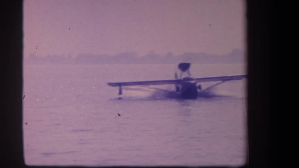 1959 8mm frame of a small float plane appearing slightly tilted or angled while moving across calm water.