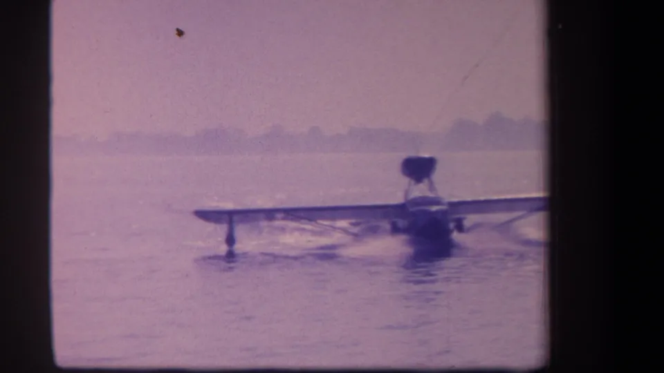 1959 8mm frame of an amphibian aircraft taxiing on calm water, with wings spread wide and a hazy shoreline in the distance.