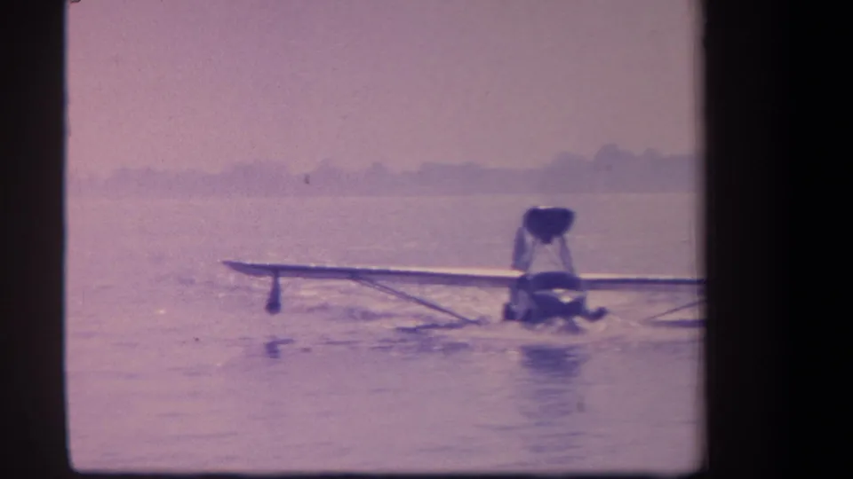 The amphibian aircraft continues across the water in this 1959 home-movie frame, leaving a light wake under a hazy sky.