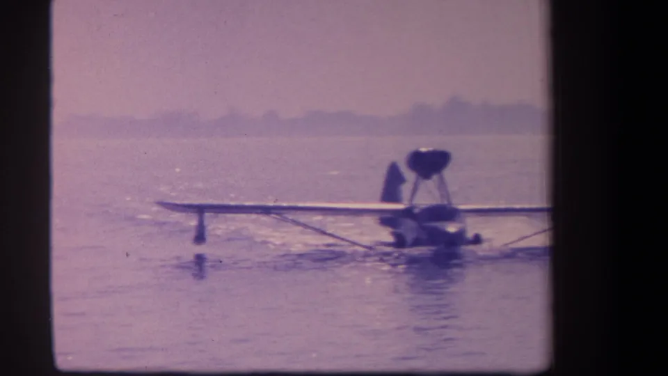 A mid-century amphibian aircraft appears in soft 8mm color as it taxis on calm water near a faint shoreline.