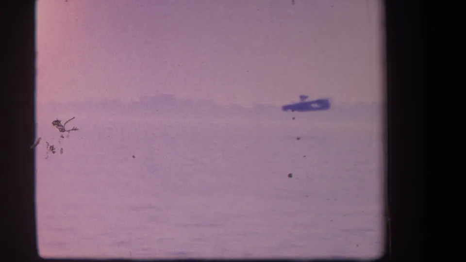 The amphibian aircraft recedes across the water in the final 1959 frame, becoming small against the hazy shoreline and purple film cast.