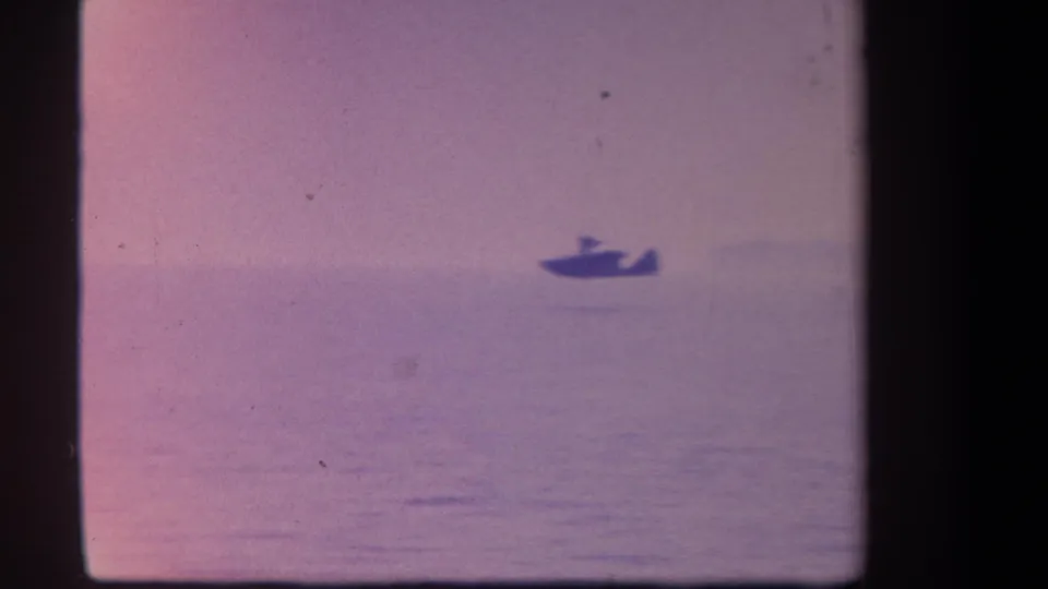 Distant hydroplane-like silhouette on open water in a faded 1959 8mm home-movie frame, with dark film borders visible.