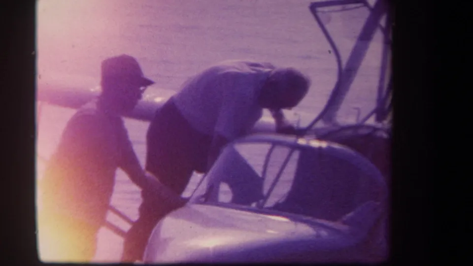 Two men work beside part of a small hydroplane or watercraft near shore in a faded 1959 8mm home-movie frame.