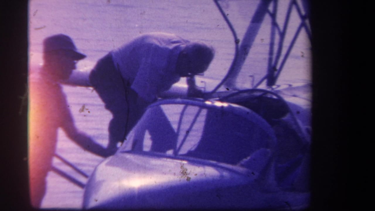 Two men beside a small float-equipped plane on a lake, one leaning into the cockpit.