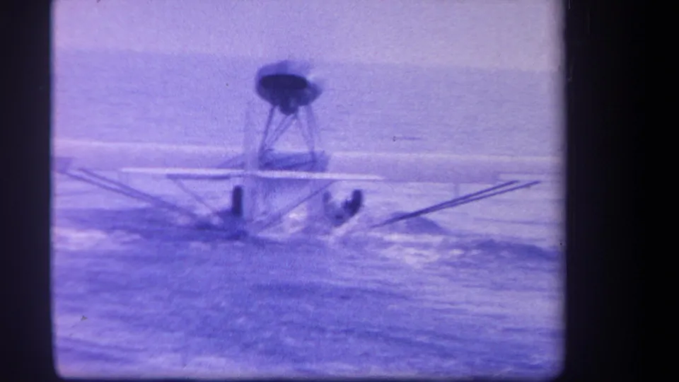 The float-equipped plane travels across the Yucaipa lake surface in faded 8mm footage, leaving wake and spray as it attempts takeoff.