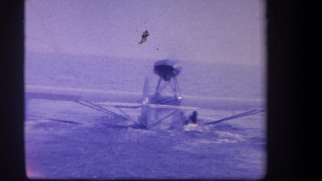 1959 8mm frame of a small seaplane or floatplane on water, seen at distance with a purple color cast and indistinct people near the fuselage.