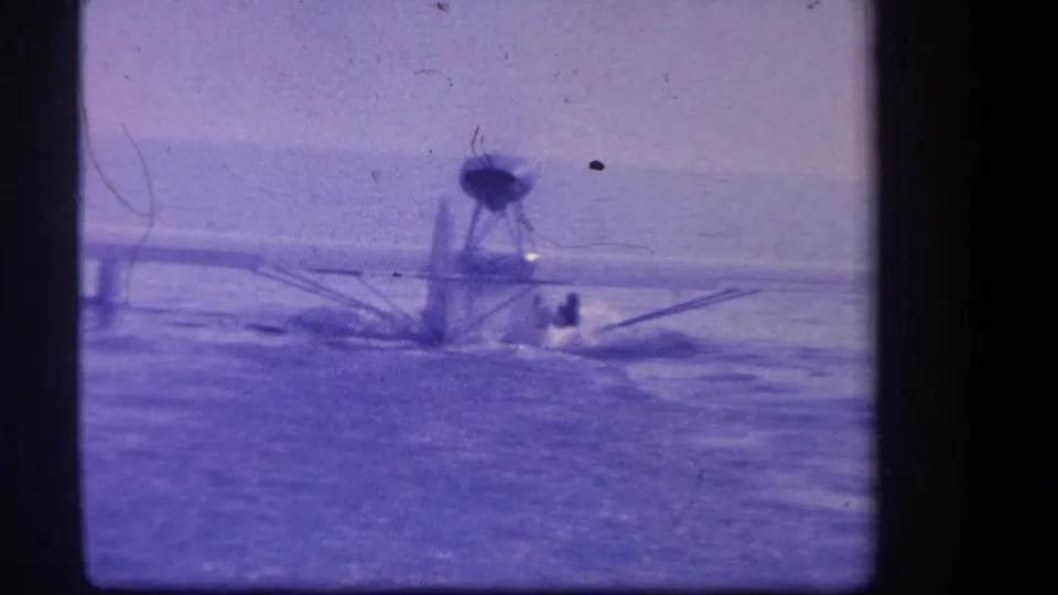 Purple-tinted 1959 home-movie frame showing a small seaplane on water, with soft focus, visible grain, and indistinct people near the aircraft.