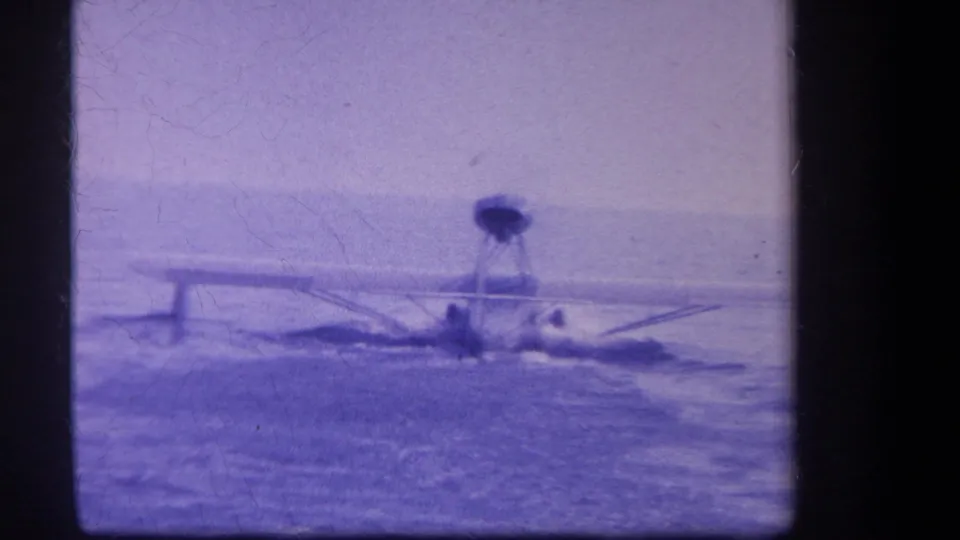 End frame of a small seaplane or floatplane floating on water in 1959 8mm footage, with purple cast, soft detail, and distant figures visible.