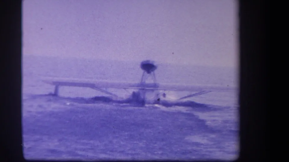1959 8mm frame from Yucaipa, California, showing a small seaplane low on the lake surface with ripples spreading behind it.