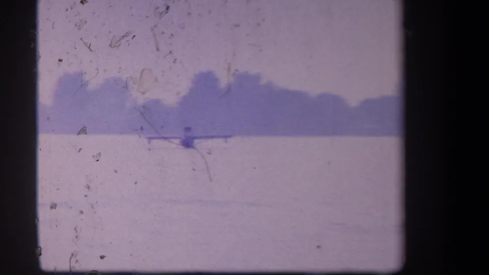 Distant view of a small seaplane on a calm Yucaipa lake, with a hazy treeline and visible 8mm scratches in the frame.