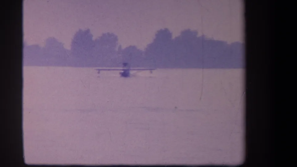 Small seaplane viewed from behind on pale lake water in the supplied 1959 Yucaipa home-movie footage, with trees beyond the shoreline.