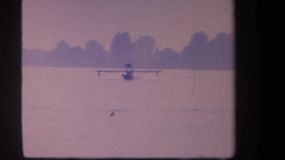 1959 8mm lake scene with a small seaplane centered on the water, faint wake marks, a distant treeline, and a purple color cast.