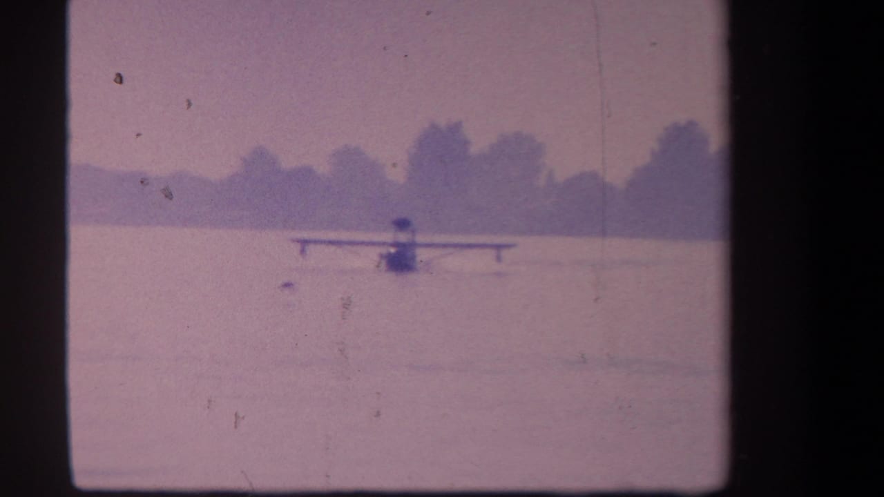 1959 8mm frame from Volmer Yucaipa showing a distant floatplane on a calm lake with a soft tree-lined horizon.