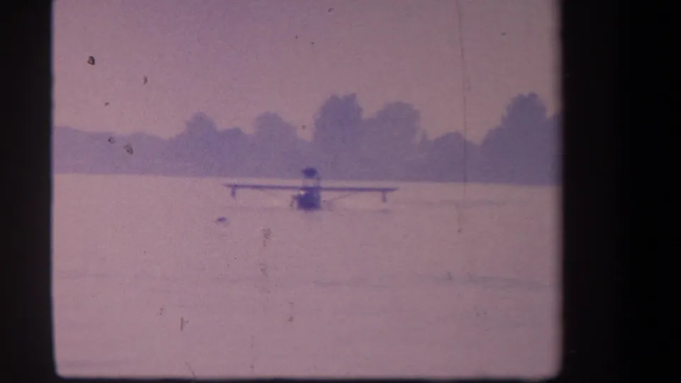 1959 8mm frame from Volmer Yucaipa showing a distant floatplane on a calm lake with a soft tree-lined horizon.