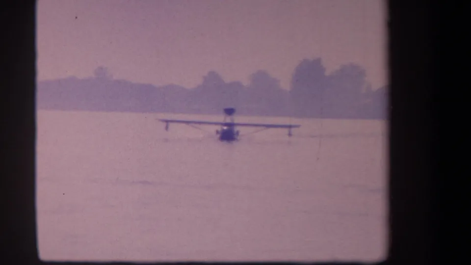1959 Volmer Yucaipa home-movie frame with a single-prop floatplane taxiing across a quiet lake before a hazy shoreline.