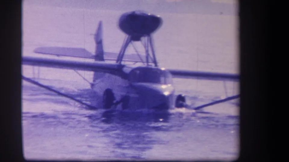 Front-facing 1959 8mm view of a likely Volmer Yucaipa amphibious aircraft floating with wing struts spread across the water.