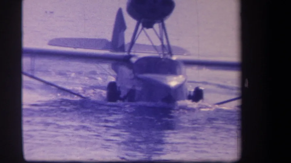 1959 home-movie frame showing the amphibious aircraft nearly head-on, its fuselage low in the water beneath a raised propeller pod.
