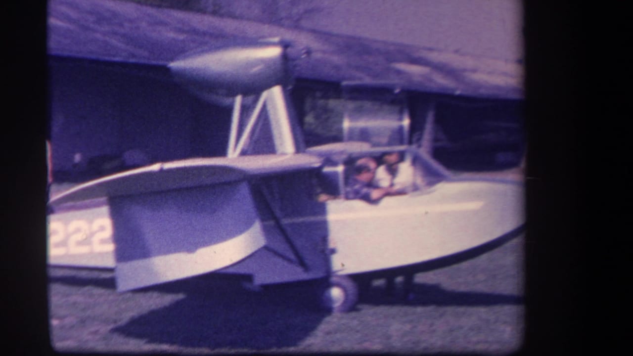 1959 Yucaipa home-movie frame showing a silver amphibious aircraft on grass, with two adult men visible at the cockpit area.