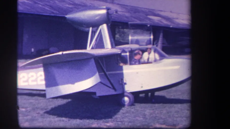 1959 home-movie frame of the amphibious aircraft on grass, emphasizing the wing, struts, cockpit, and two men inside or beside the cabin.