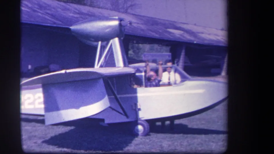 1959 Yucaipa frame with occupants in the cockpit of a high-wing amphibious aircraft parked near a building.