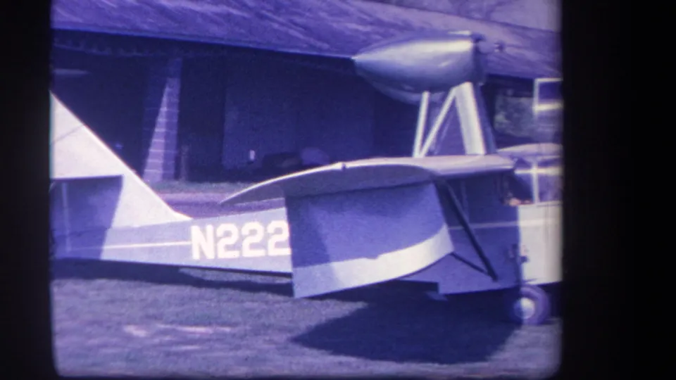 1959 8mm frame of the aircraft side, showing part of registration N222, high wing, landing wheel, and shaded building behind.