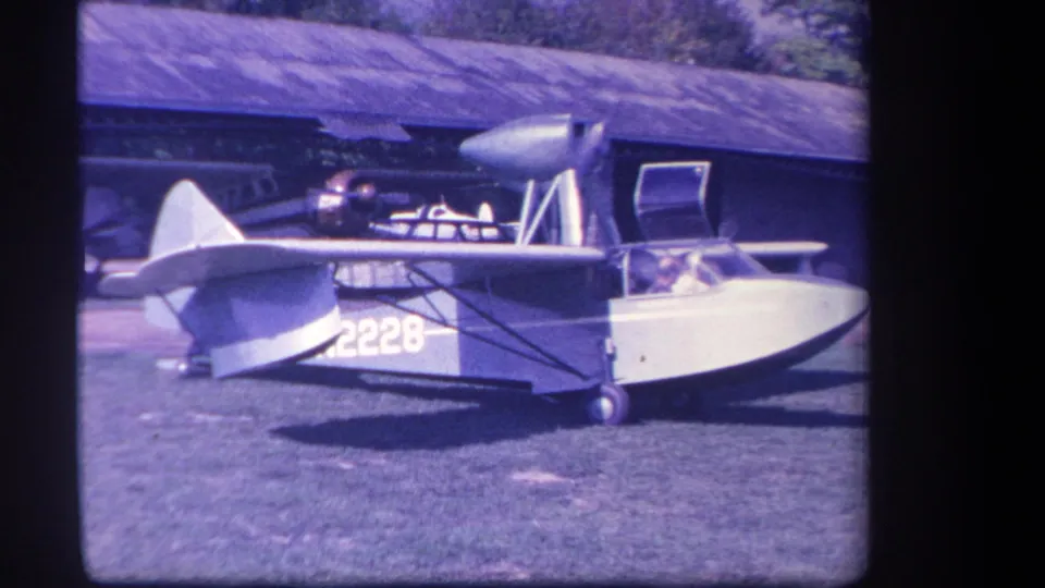 1959 home-movie frame of amphibious plane N2228 on grass, with its open cockpit and rural barn-like backdrop visible.
