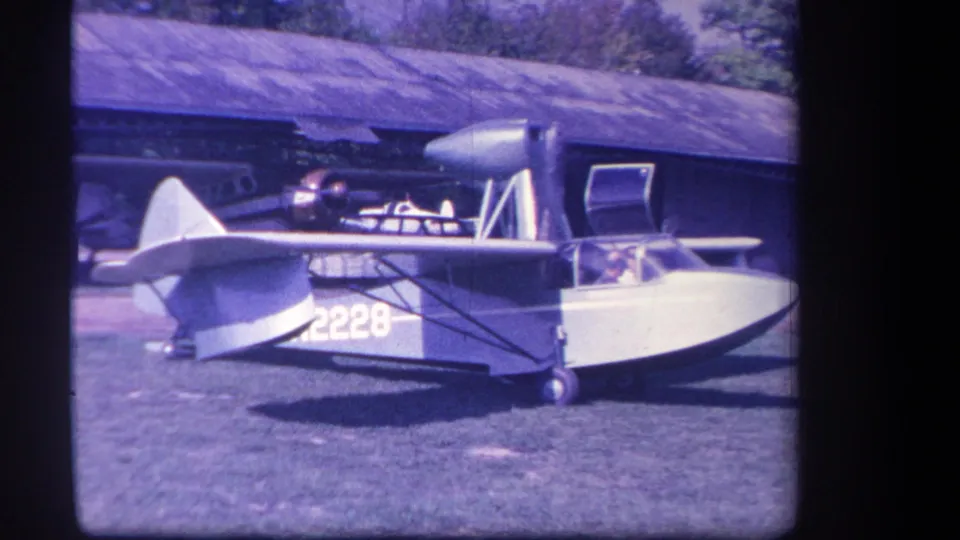 1959 home-movie frame from Yucaipa showing a small amphibious aircraft on grass with a cockpit occupant and tail number 2228 visible.