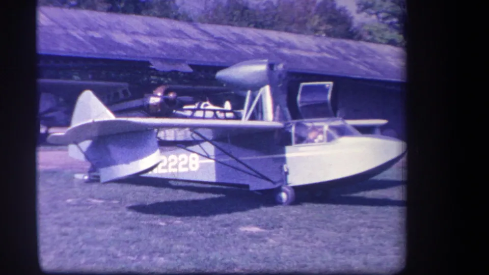 1959 Yucaipa frame showing the amphibious airplane in profile, with its boat-like hull, cockpit area, propeller, and background building.