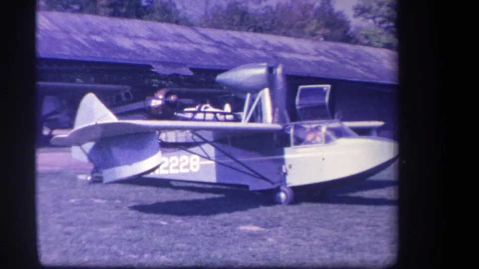 Vintage 8mm frame from the Yucaipa clip showing the stationary amphibious aircraft, cockpit occupant, struts, and low building in the background.