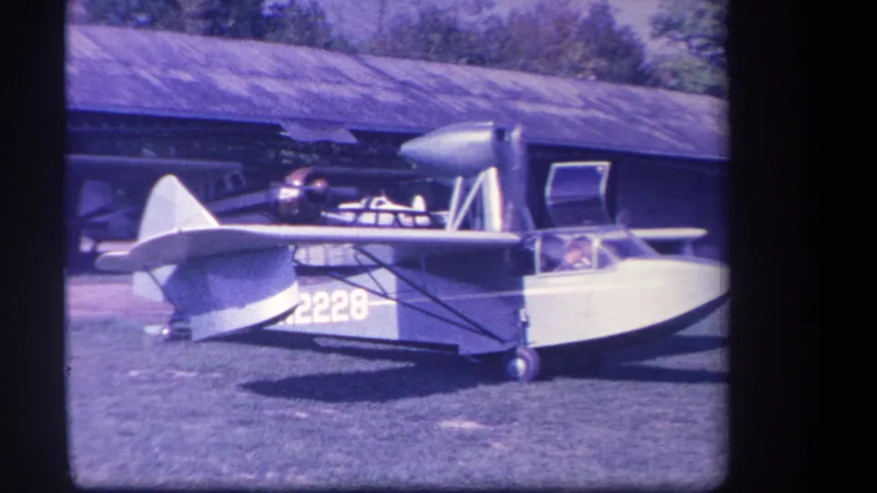 A closer 1959 frame shows the amphibious plane's side, wing struts, number 2228, cockpit area, and open door against a hangar backdrop.