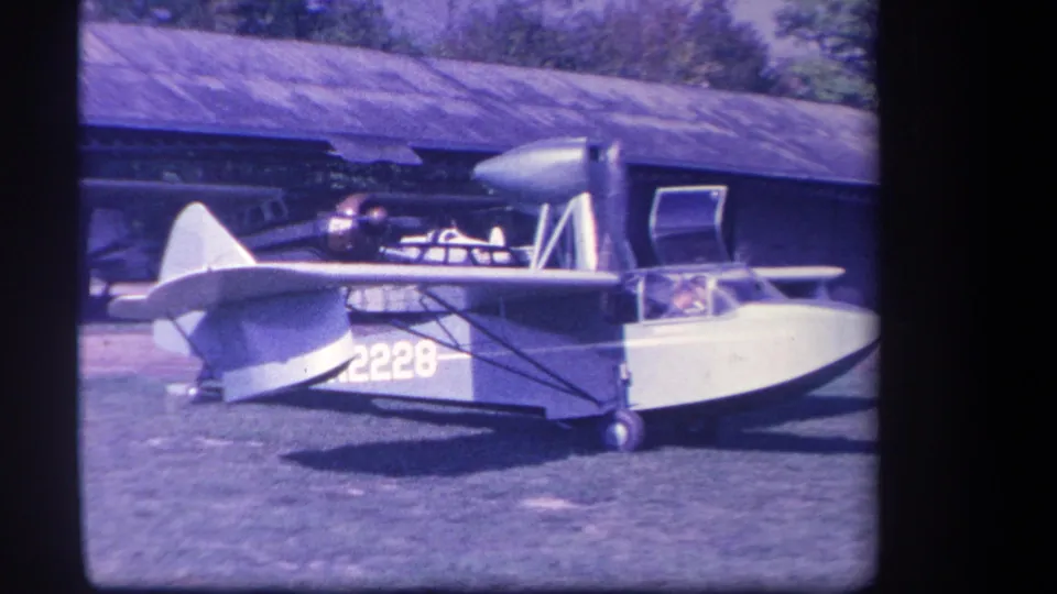 The closing frame keeps the amphibious aircraft centered on the grass field, with propeller blur, open door, cockpit figure, and hangar structure visible.