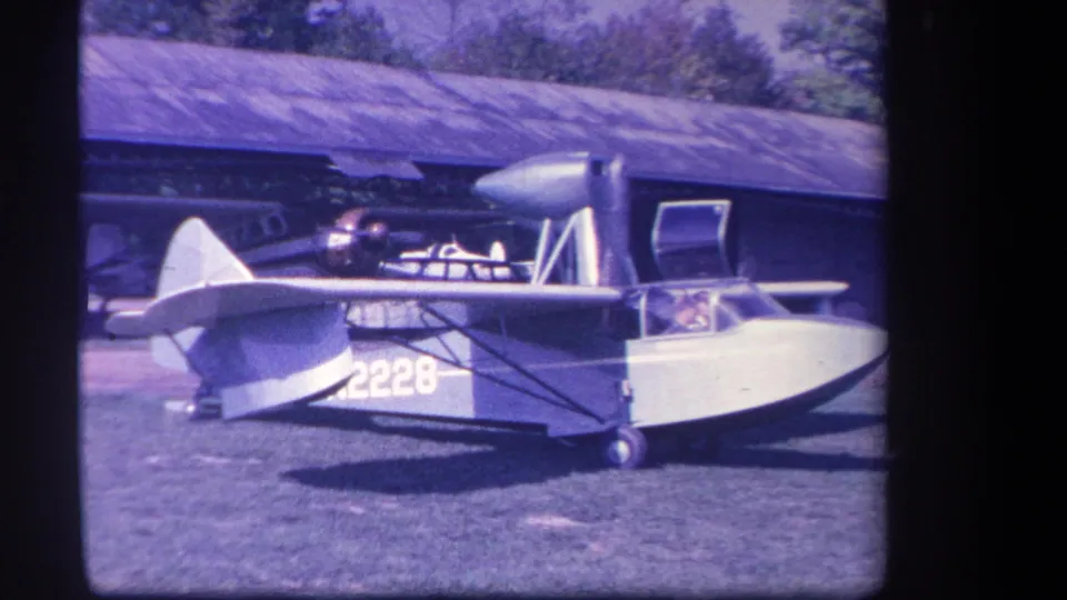 1959 Yucaipa-area home-movie frame showing a small blue and white amphibious aircraft with number 2228 beside a hangar.