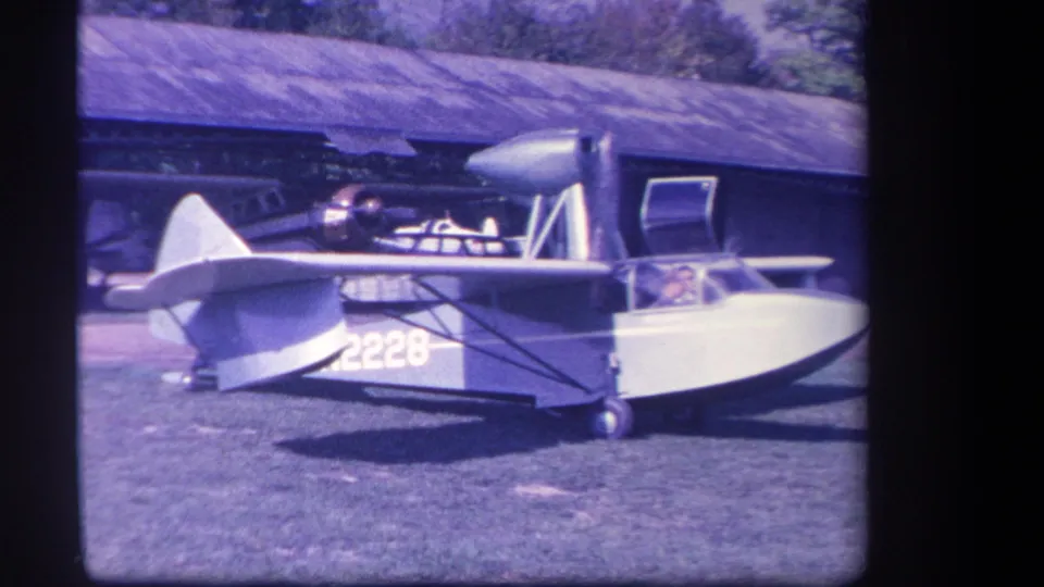 1959 Yucaipa-area frame showing the amphibious aircraft from the side, with a person visible in the cockpit and hangar structure behind.