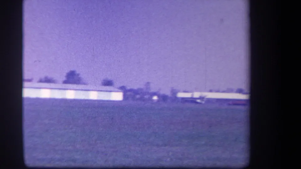 Wide 1959 Yucaipa-area home-movie view of the airfield, with open grass, low hangar buildings, distant trees, and pale sky.