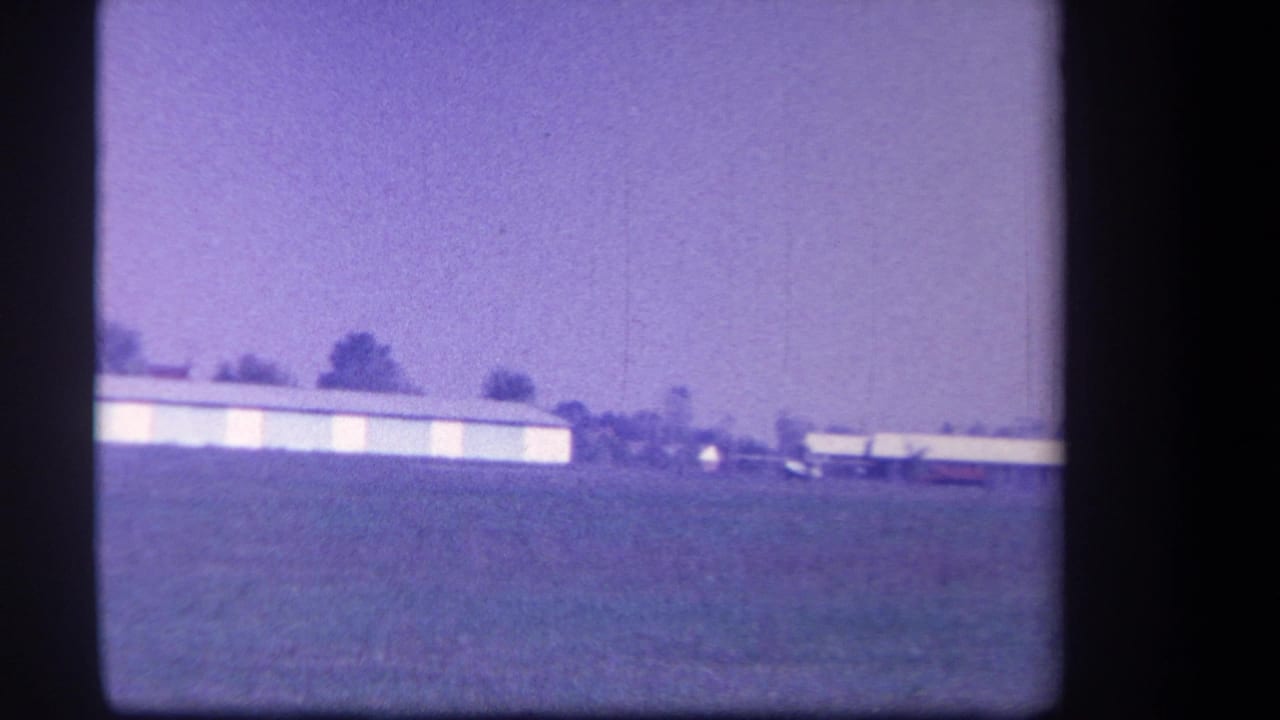 Distant hangar and small plane across a grass field under a hazy sky.