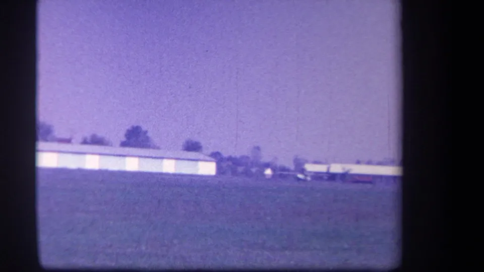 1959 Yucaipa-area home-movie frame showing a low hangar and small aircraft viewed from across a grass field.