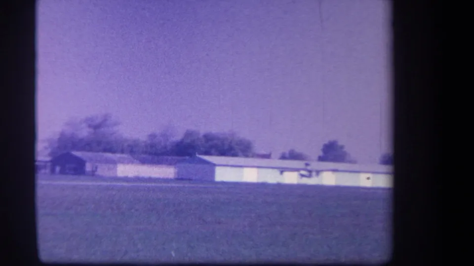 Soft 8mm view of the 1959 Yucaipa-area airfield with low buildings, trees, and open grass in the foreground.
