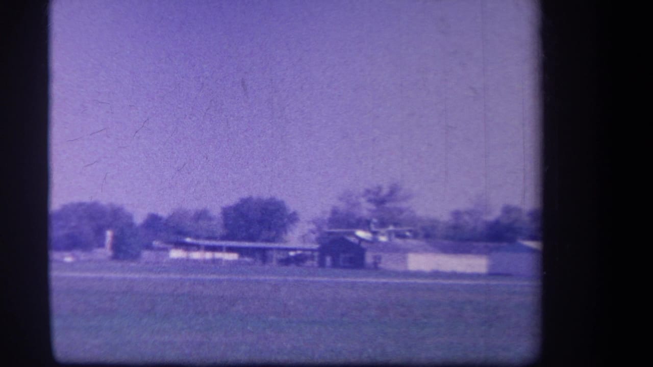 1959 8mm frame from Yucaipa area showing a distant rural airfield with grass, low structures, trees, and aged purple color shift.