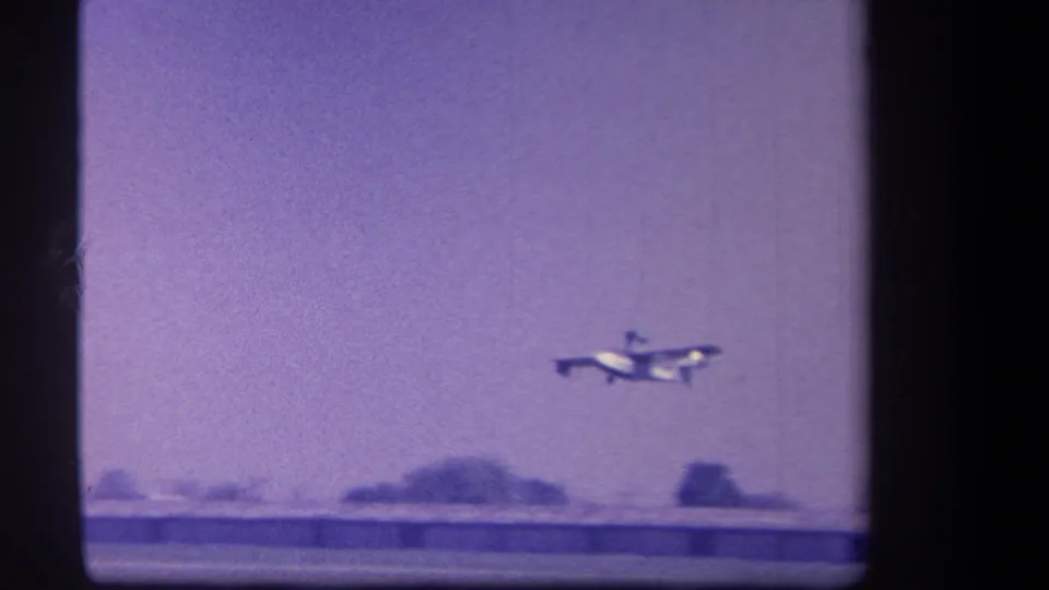 The propeller plane lifts into the air over the rural airfield, with low structures and a blurred tree line below.